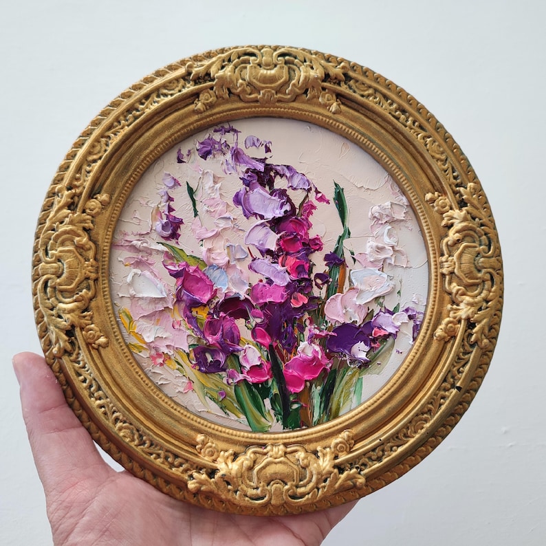 May include: A circular oil painting featuring thick impasto brushstrokes of purple, magenta, and white flowers with green stems. The artwork is encased in an ornate, circular gold-colored frame with intricate relief detailing along the border.