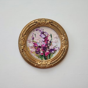 May include: A circular oil painting featuring thick, textured impasto brushstrokes of purple, pink, and white flowers with green stems. The artwork is encased in an ornate, circular gold frame with intricate relief detailing along the edges.