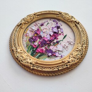 May include: A circular oil painting featuring thick, textured impasto brushstrokes of purple, pink, and white flowers with green stems. The artwork is encased in an ornate, round, metallic gold frame with intricate embossed scrollwork details.