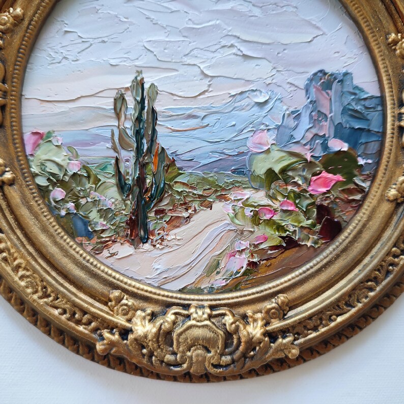 May include: An oval oil painting featuring a desert landscape with a tall cactus, rocky formations, and a winding path, rendered with thick impasto brushstrokes in shades of blue, green, and pink, enclosed in an ornate, antique gold-toned frame.