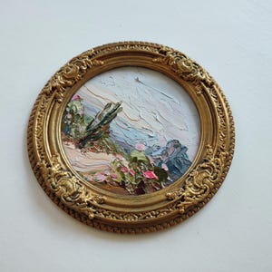 May include: A circular oil painting featuring a desert landscape with cacti and pink flowers, rendered with thick, textured brushstrokes. The artwork is encased in an ornate, antique-style gold frame with intricate scrollwork and relief detailing.