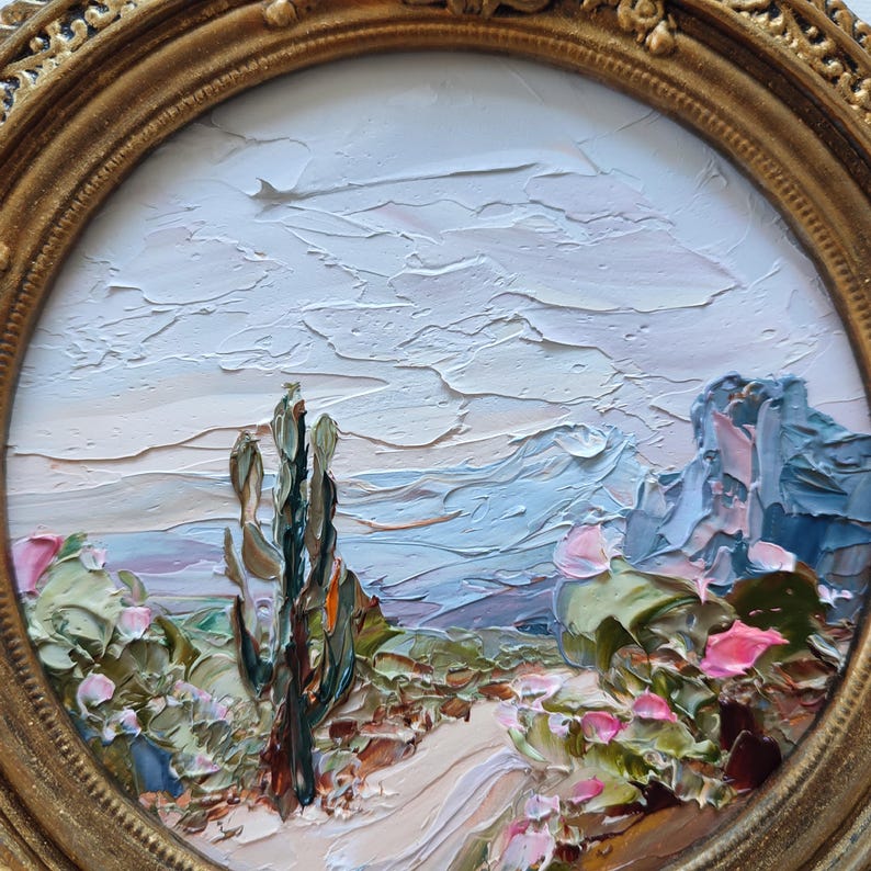 May include: An oil painting in a round gold frame depicting a desert landscape with a tall central cactus, low-lying prickly pear cacti with pink flowers, a winding path, and distant blue mountains under a textured, pale, cloudy sky.