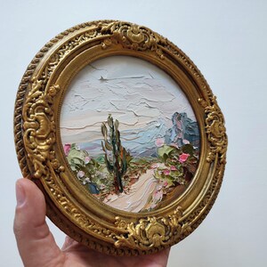 May include: A circular oil painting featuring a textured landscape with a central cactus, a winding path, and distant mountains under a pale sky. The artwork is encased in an ornate, antique-style gold frame with intricate relief detailing.