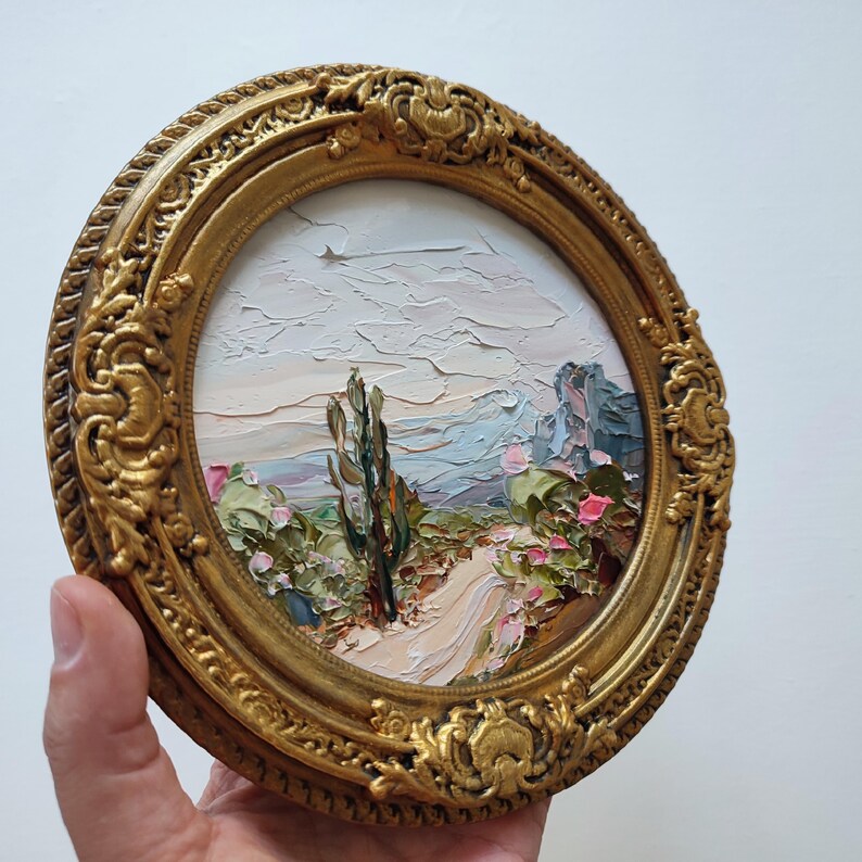 May include: A circular oil painting featuring a textured landscape with a central cactus, a winding path, and distant mountains under a pale sky. The artwork is encased in an ornate, antique-style gold frame with intricate relief detailing.