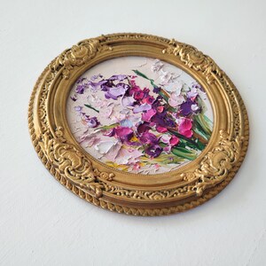 May include: A circular oil painting featuring thick, textured impasto brushstrokes of purple, magenta, and white flowers. The artwork is encased in an ornate, round, metallic gold frame with intricate embossed detailing along the outer edge.