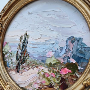 May include: An oil painting in a round, ornate gold frame depicting a desert landscape with a tall, dark cactus, green succulents, and bright pink flowers. The scene features thick, textured brushstrokes and a pale, cloudy sky above mountains.
