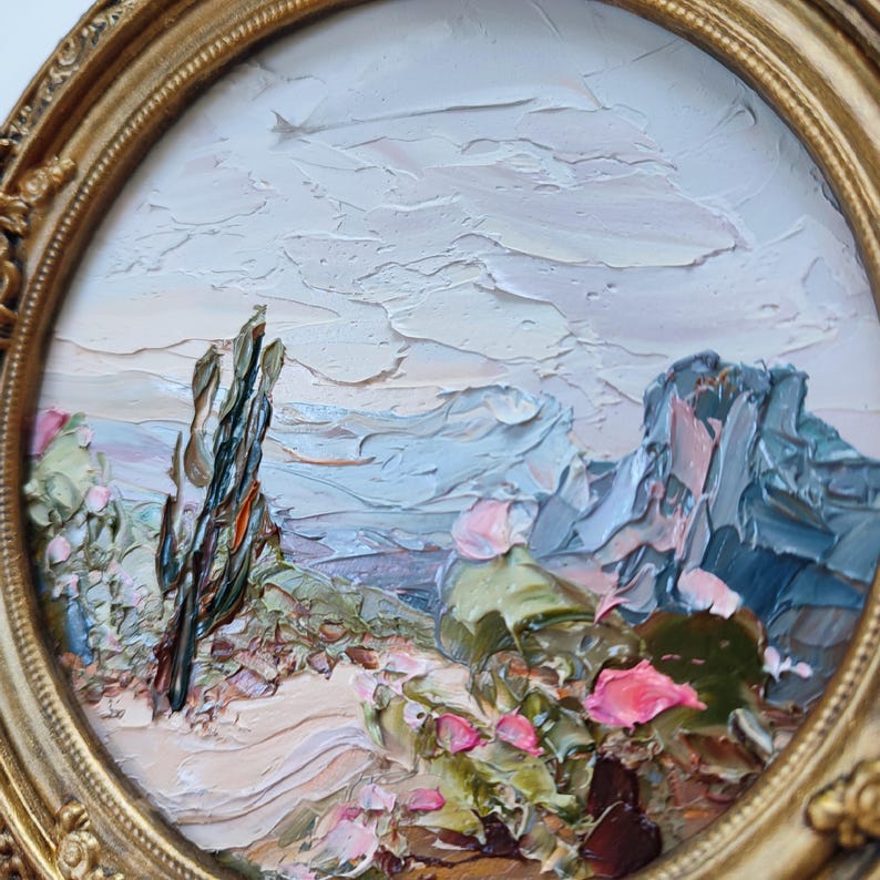 May include: An oil painting in a round, ornate gold frame depicting a desert landscape with a tall, dark cactus, green succulents, and bright pink flowers. The scene features thick, textured brushstrokes and a pale, cloudy sky above mountains.