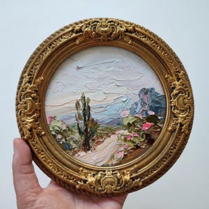 May include: A circular oil painting featuring a textured landscape with a central cactus, a winding path, and distant mountains under a pale sky, encased in an ornate, antique-style gold frame with intricate scrollwork and decorative relief details.