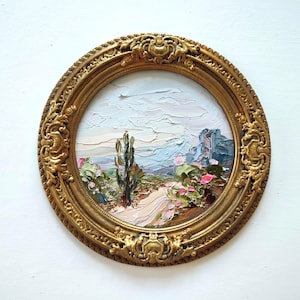 May include: A circular oil painting featuring a landscape with a central tall cactus, a winding path, and distant mountains under a pale sky. The artwork is encased in an ornate, decorative gold-colored frame with intricate relief detailing.