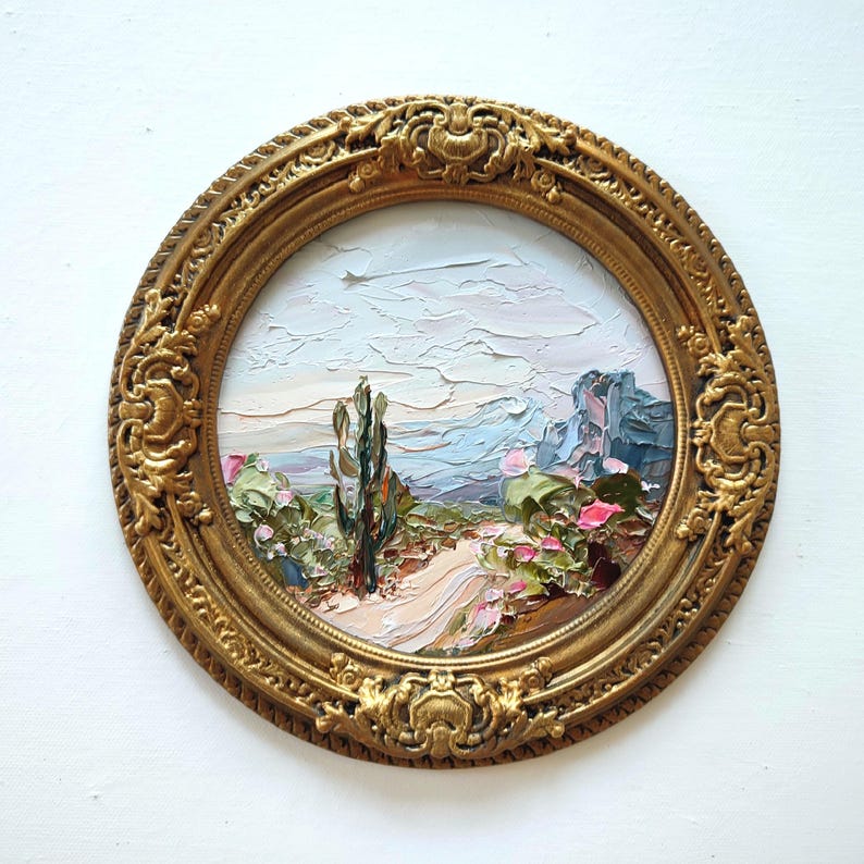May include: A circular oil painting featuring a landscape with a central tall cactus, a winding path, and distant mountains under a pale sky. The artwork is encased in an ornate, decorative gold-colored frame with intricate relief detailing.