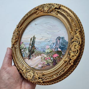 May include: A round oil painting in a decorative gold frame featuring a textured landscape with a tall thin tree, rocky formations, and pink floral accents under a pale sky. The artwork uses thick impasto brushstrokes to create depth.