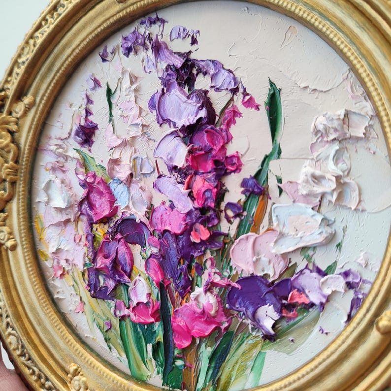 May include: An oil painting in a round, ornate gold frame featuring a thick impasto floral arrangement. The artwork displays vibrant shades of purple, magenta, and pink flowers with green stems against a pale, textured off-white background.