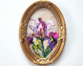 May include: An oval oil painting featuring a textured impasto iris flower in shades of purple, pink, and yellow with green leaves. The artwork is encased in an ornate, decorative gold-coloured frame with intricate relief detailing.