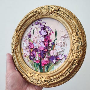 May include: A circular oil painting featuring thick, textured impasto brushstrokes of purple, magenta, and white flowers with green stems. The artwork is encased in an ornate, circular gold-toned frame with intricate relief detailing.