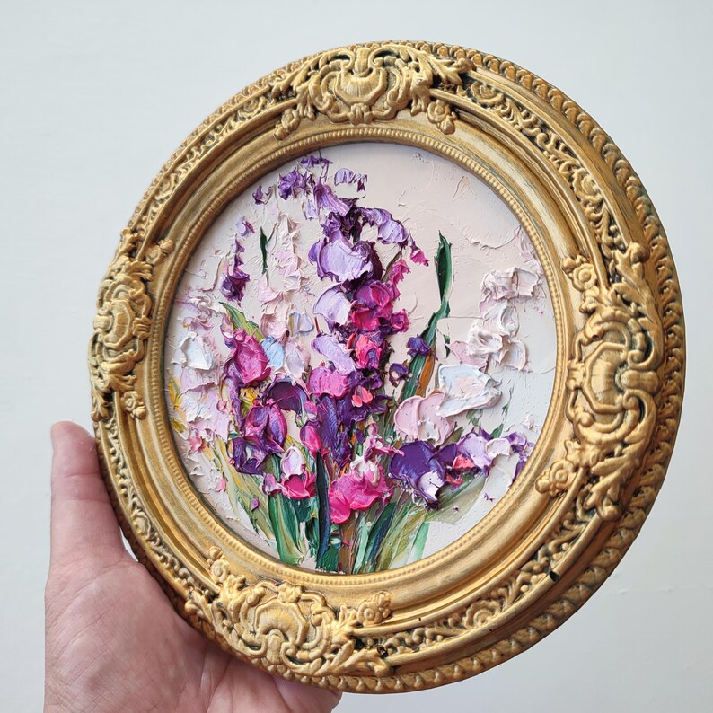 May include: A circular oil painting featuring thick, textured impasto brushstrokes of purple, magenta, and white flowers with green stems. The artwork is encased in an ornate, circular gold-toned frame with intricate relief detailing.