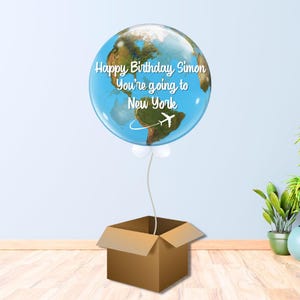 May include: A clear balloon, printed with a world map and the text "Happy Birthday Simon You're going to New York", floats above a brown cardboard box. A white airplane graphic is also on the balloon.