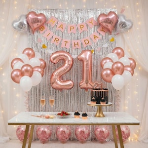 Może przedstawiać: Uroczysta scena urodzinowa z białym marmurowym stołem, ozdobionym balonami, tortem i kieliszkami do szampana. Balony w kolorze różowego złota z liczbą "21", z banerem "Happy Birthday". Tło to srebrna frędzla.