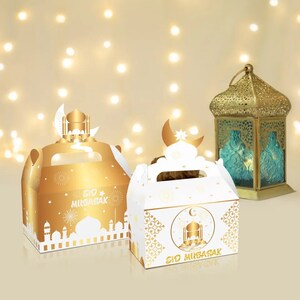 May include: Two gold and white gift boxes with handles, decorated with a crescent moon and mosque design. The boxes have the text "Eid Mubarak" on them. The boxes are in front of a gold lantern with a teal glass panel.