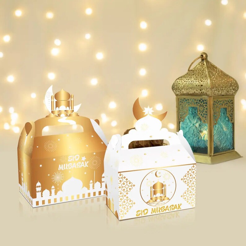 May include: Two gold and white gift boxes with handles, decorated with a crescent moon and mosque design. The boxes have the text "Eid Mubarak" on them. The boxes are in front of a gold lantern with a teal glass panel.