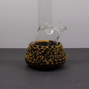 May include: A clear glass bong with a green rim and a black and gold crocheted cozy.