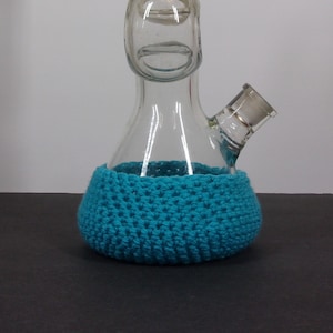 May include: A clear glass bong with a blue crocheted base. The bong has a curved neck and a round chamber.