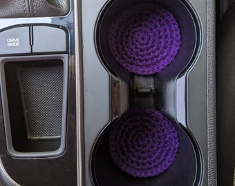 May include: Two purple crocheted cup holders in a car's center console. The console has a black finish and a silver trim. The cup holders are in the lower right corner of the image. The console also has a drive mode button.