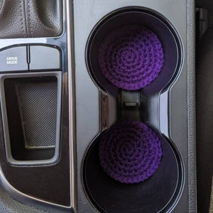 May include: Two purple crocheted cup holders in a car's center console. The console has a black finish and a silver trim. The cup holders are in the lower right corner of the image. The console also has a drive mode button.