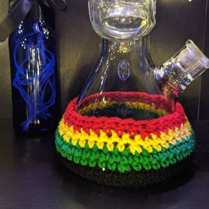 May include: A glass bong with a crocheted cover in the colors of the Jamaican flag. The cover is red, yellow, and green. The bong has a round base and a long neck.
