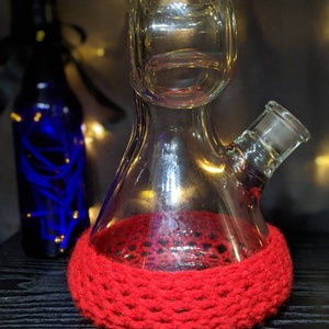 May include: A clear glass bong with a red crocheted base. The bong has a round chamber and a small, round mouthpiece.