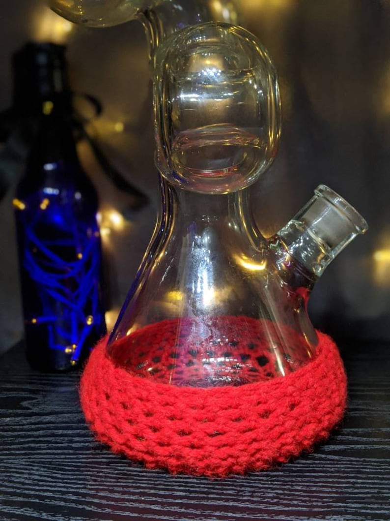 May include: A clear glass bong with a red crocheted base. The bong has a round chamber and a small, round mouthpiece.