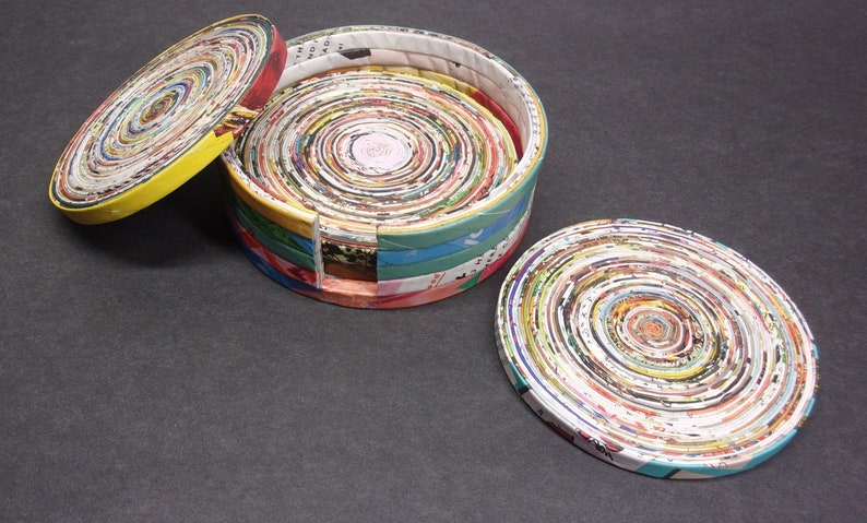 May include: A round, colorful box made from rolled up magazine pages. The box is open and the lid is laying next to it. The box and lid are both decorated with a spiral pattern of colorful magazine pages.