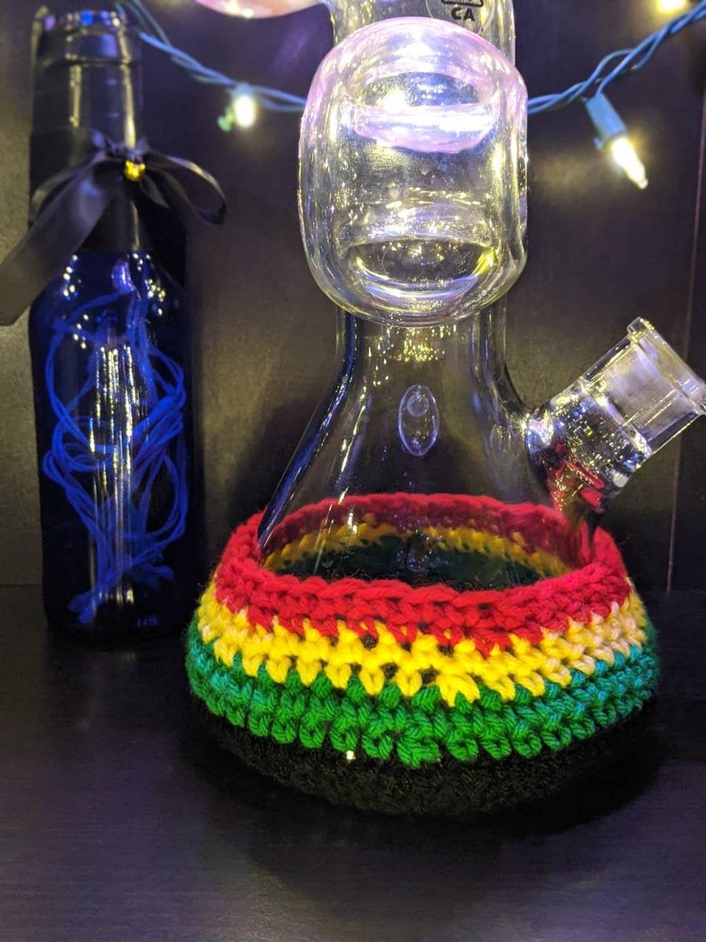 May include: A glass bong with a crocheted cover in the colors of the Jamaican flag. The cover is red, yellow, and green. The bong has a round base and a long neck.