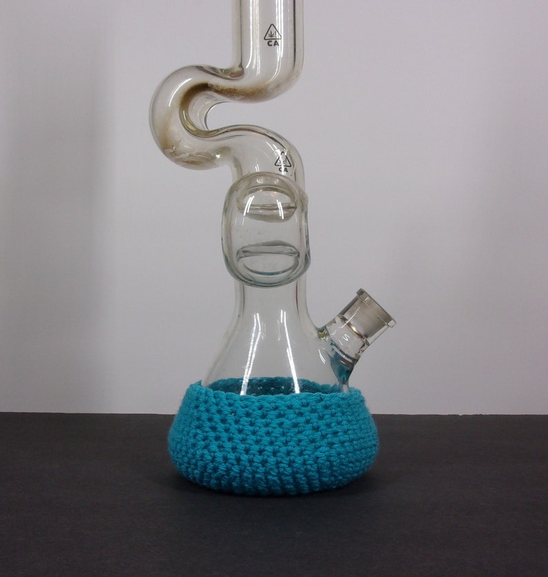May include: A clear glass bong with a blue crocheted base. The bong has a curved neck and a round chamber.