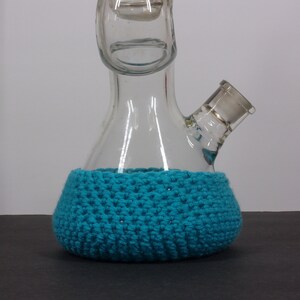 May include: A clear glass bong with a blue crocheted base. The bong has a round base and a long neck with a small opening at the top.