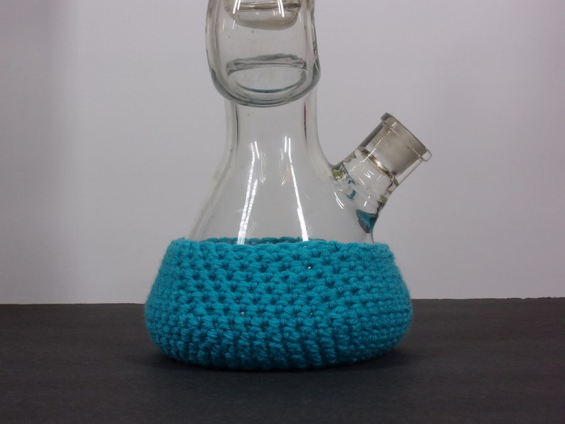 May include: A clear glass bong with a blue crocheted base. The bong has a round base and a long neck with a small opening at the top.