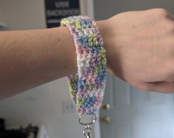 May include: A crocheted wristlet with a light blue, pink, green, and white color pattern. The wristlet has a silver key chain attached.