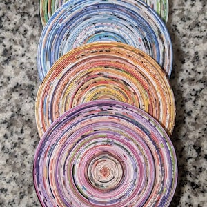 May include: Four round coasters made from rolled up magazine pages. The coasters are stacked on top of each other. The top coaster is purple, the next is orange, the next is blue, and the bottom coaster is green.