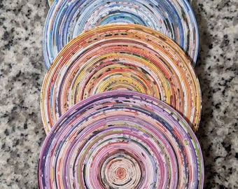 May include: Four round coasters made from rolled up magazine pages. The coasters are stacked on top of each other. The top coaster is purple, the next is orange, the next is blue, and the bottom coaster is green.