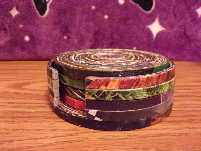May include: A stack of round coasters made from rolled up magazine pages. The coasters are in various colors and patterns, including black, green, red, and brown.
