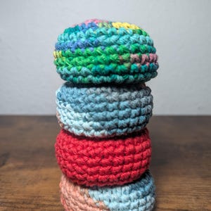 May include: A stack of four crocheted circular items in various colors. The top item is a mix of green, blue, and yellow. Below it is a blue item, followed by a red one, and the bottom item is pink and blue. The items are stacked on a wooden surface.