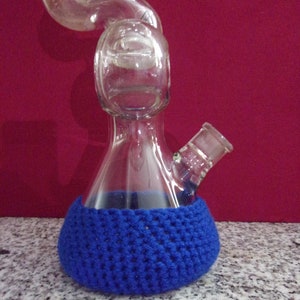 May include: A clear glass beaker with a curved neck and a blue crocheted cover. The beaker has a small spout on the side.