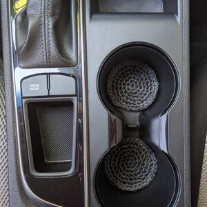 May include: Two black cup holders with gray crocheted cup coasters inside. The cup holders are part of a black car interior with a gear shift lever and a "Drive Mode" button.