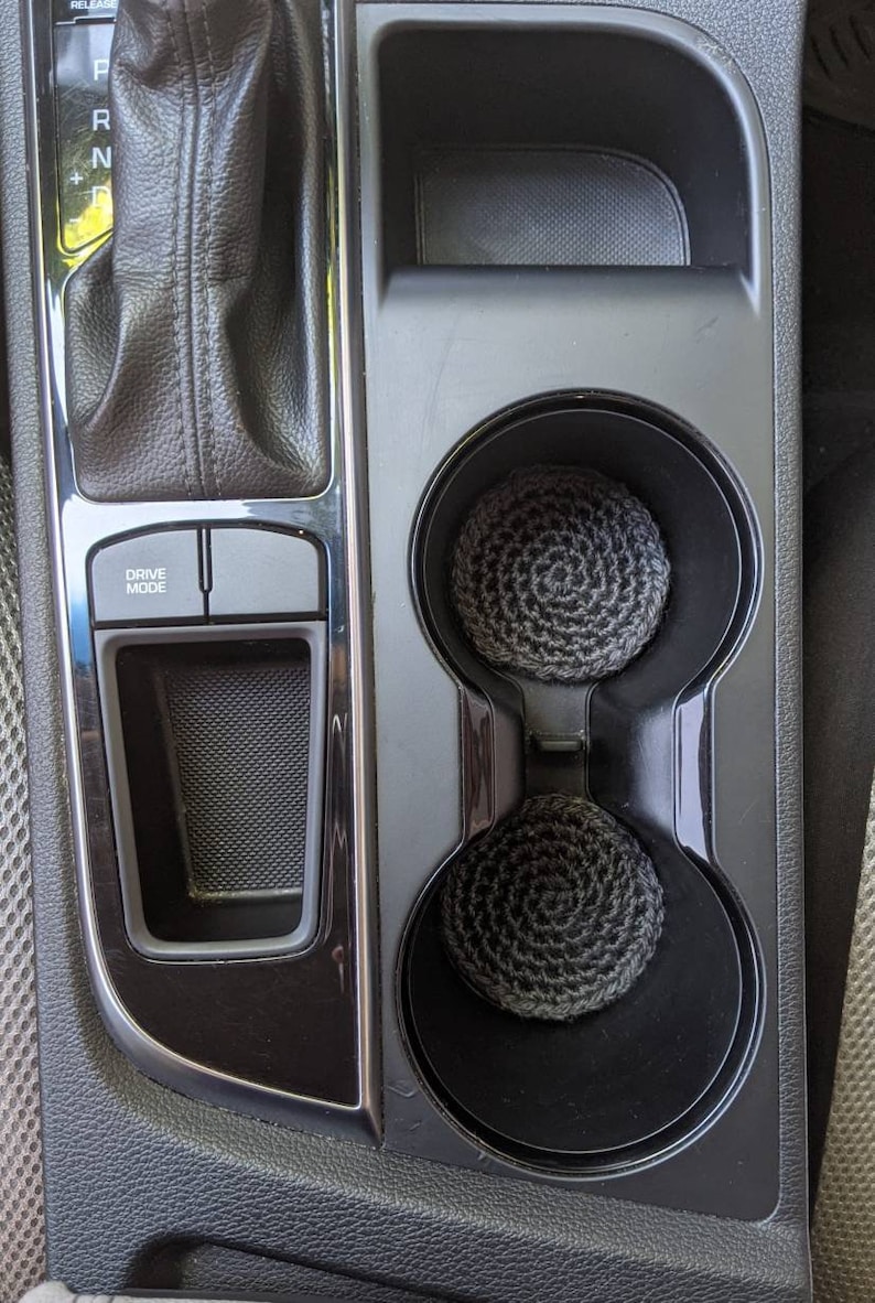 May include: Two black cup holders with gray crocheted cup coasters inside. The cup holders are part of a black car interior with a gear shift lever and a "Drive Mode" button.