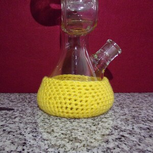 May include: A clear glass bong with a yellow crocheted cover. The bong has a round base and a long neck with a bowl on top.