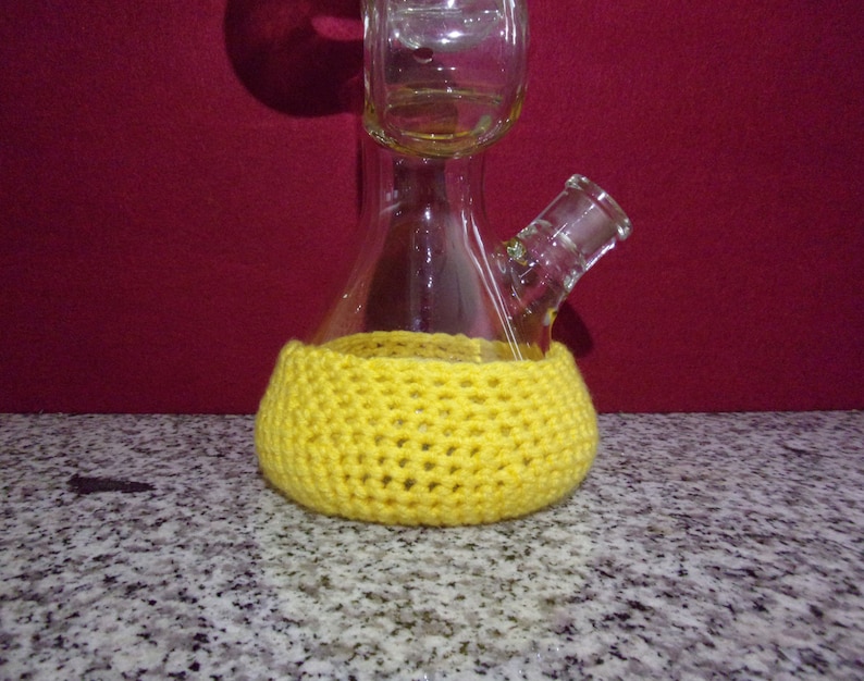 May include: A clear glass bong with a yellow crocheted cover. The bong has a round base and a long neck with a bowl on top.