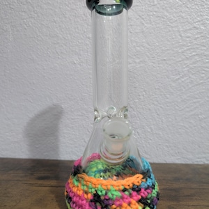 May include: A clear glass water pipe with a flared base and a dark green rim. The base is wrapped in a colorful, hand-crocheted cozy featuring neon pink, orange, green, and blue yarn. The water pipe sits on a wooden surface.