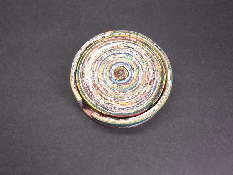 May include: A circular coaster made from rolled up magazine pages. The coaster has a concentric circle pattern with shades of green, yellow, red, and white.