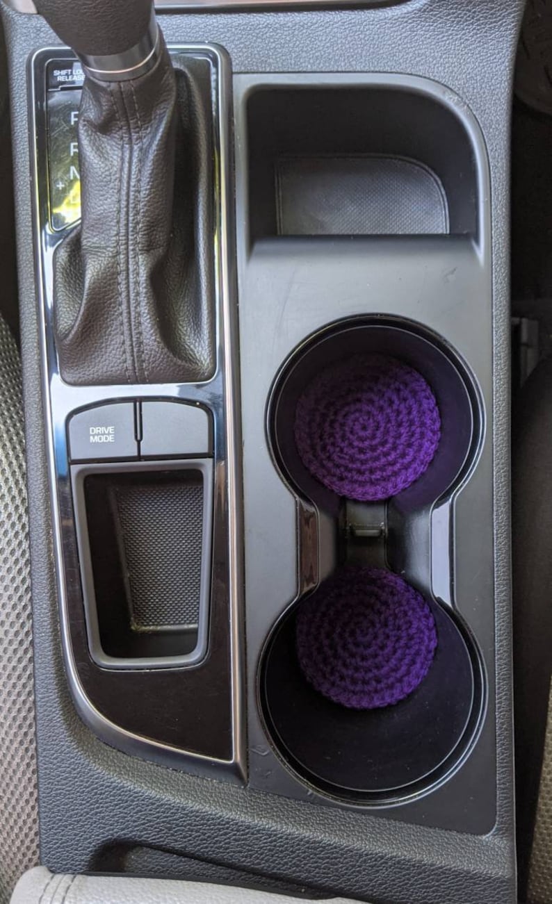May include: Two purple crocheted cup holders in a car's center console. The console has a black finish and a silver trim. The cup holders are in the lower right corner of the image. The console also has a drive mode button.