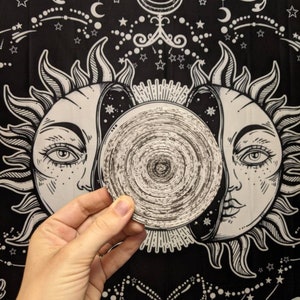 May include: A black and white tapestry with a sun and moon design. The sun and moon have faces and are surrounded by stars. A hand is holding a round, black and white object in front of the tapestry.