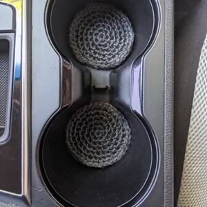 May include: Two black and gray crocheted cup holders in a car's cup holder.